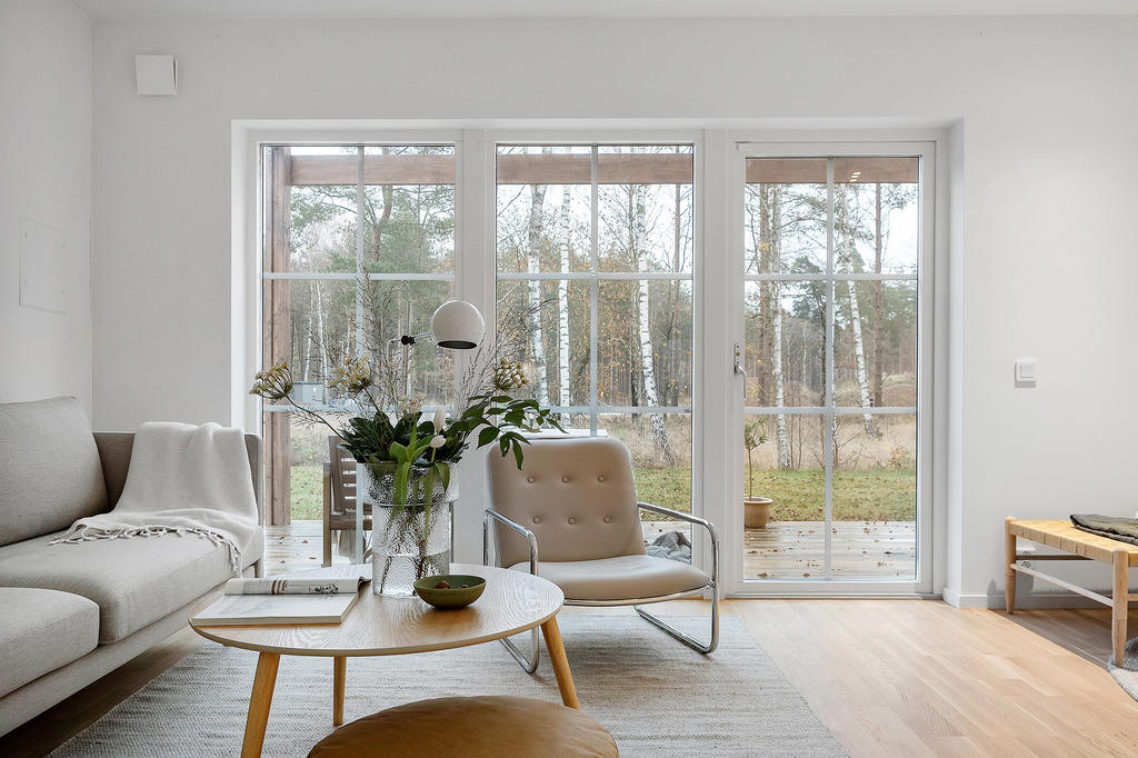 Stora fönsterpartier ner till golv i vardagsrummet ger ett härligt ljusinflöde på bostadens nedre plan.  Med uteplats och pergola precis utanför skapas en förlängd rumskänsla där du närmar dig den vackra naturen runt omkring. Inspirationsbild från visningshuset på Nässelvägen 2A (126 kvm), notera att avvikelser förekommer gällande golvet som är i ek i övriga bostäder.
