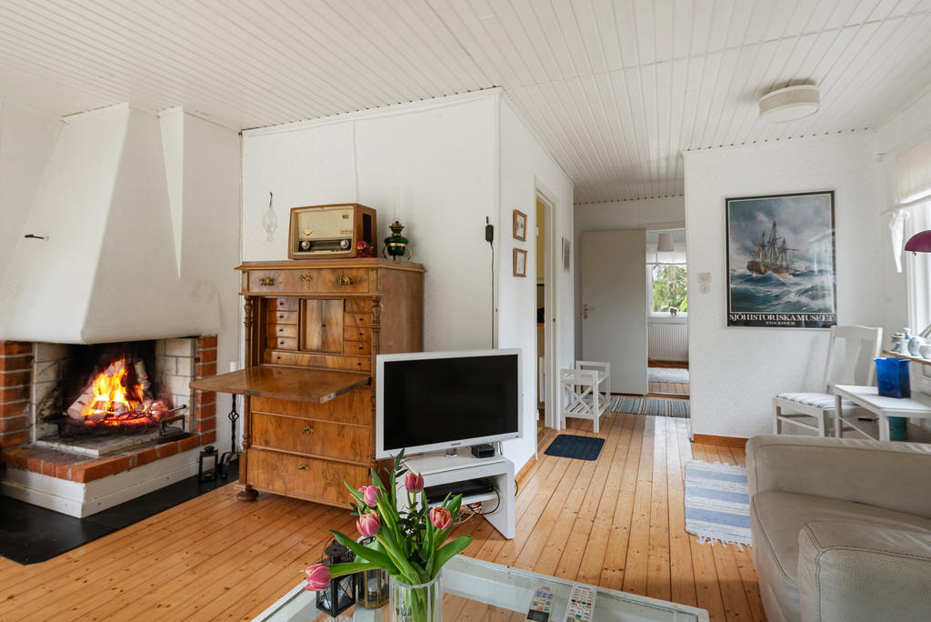 Generöst vardagsrum med stora fönster i tre väderstreck. En härlig öppen spis att mysa framför och utgång mot altanen i väster. Ljusa väggar med tapet i diskret mönster, vitt tak och det vackra trägolvet.