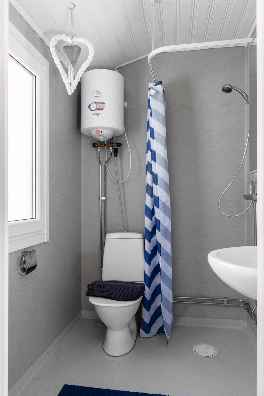 Badrum med våtrumsmatta och väggar i modern grå kulör. Utrustad med dusch, wc, handfat, varmvattensberedare och fönster