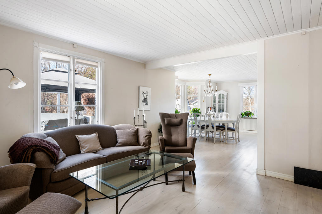Matsalen följs direkt av ett sällskapsrum med plats för en stor soffa med tillbehör. Rummet hänger ihop med matsalsdelen och har vita väggar, fönster mot uterummet i söder och mot altanen i väster. Vitpigmenterat golv och vitt tak. 