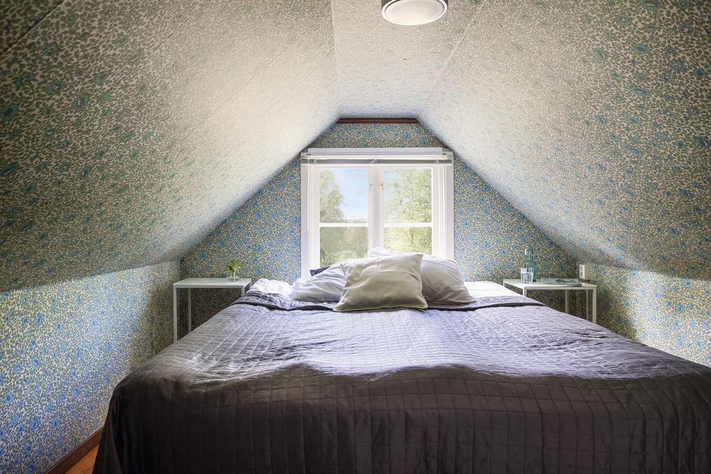 Ett charmigt loftsovrum med sluttande tak och ett centralt placerat fönster som ger fin utsikt och naturligt ljusinsläpp. Tapeten i mjuk grön nyans och låg takhöjd bidrar till en ombonad och vilsam känsla, idealiskt som gästrum eller för barn.