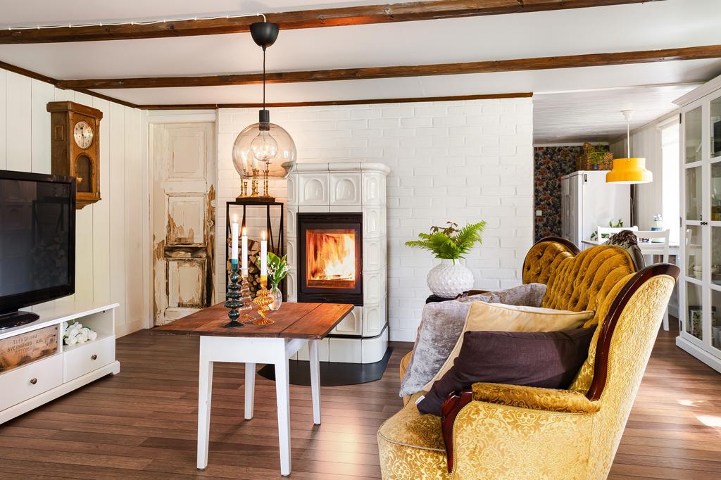 Ett luftigt och harmoniskt rum med synliga takbjälkar och ljusa väggar. Här finns både plats för soffgrupp och matplats, vilket gör rummet till en naturlig samlingspunkt. 
Kamin i gammal stil ger en behaglig värme och mysfaktor. Inredningen balanserar modern komfort med rustik charm.