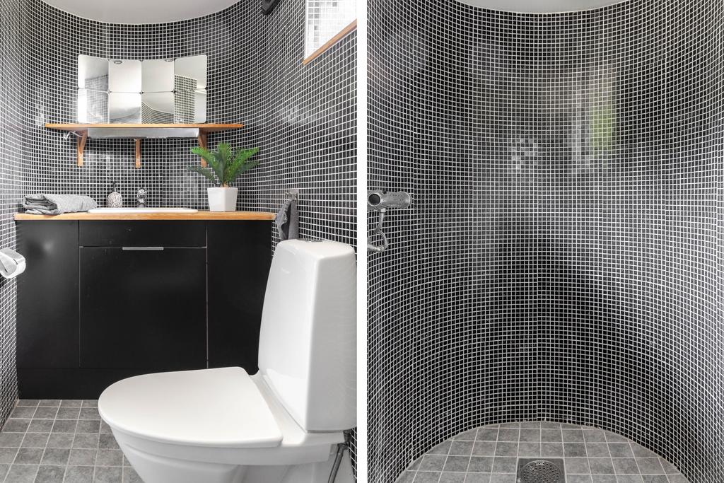 Stilrent badrum på nedre plan med wc, handfat och dusch bakom rundad vägg. Svart klinker och mosaik ger en modern känsla med karaktär.
