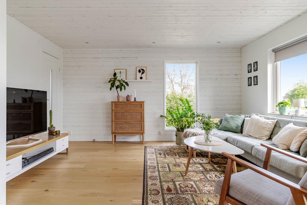 Luftigt vardagsrum med plats för både soffgrupp och tv-möbel. De stora fönstren släpper in gott om dagsljus och bjuder på utsikt mot grönskande omgivningar.