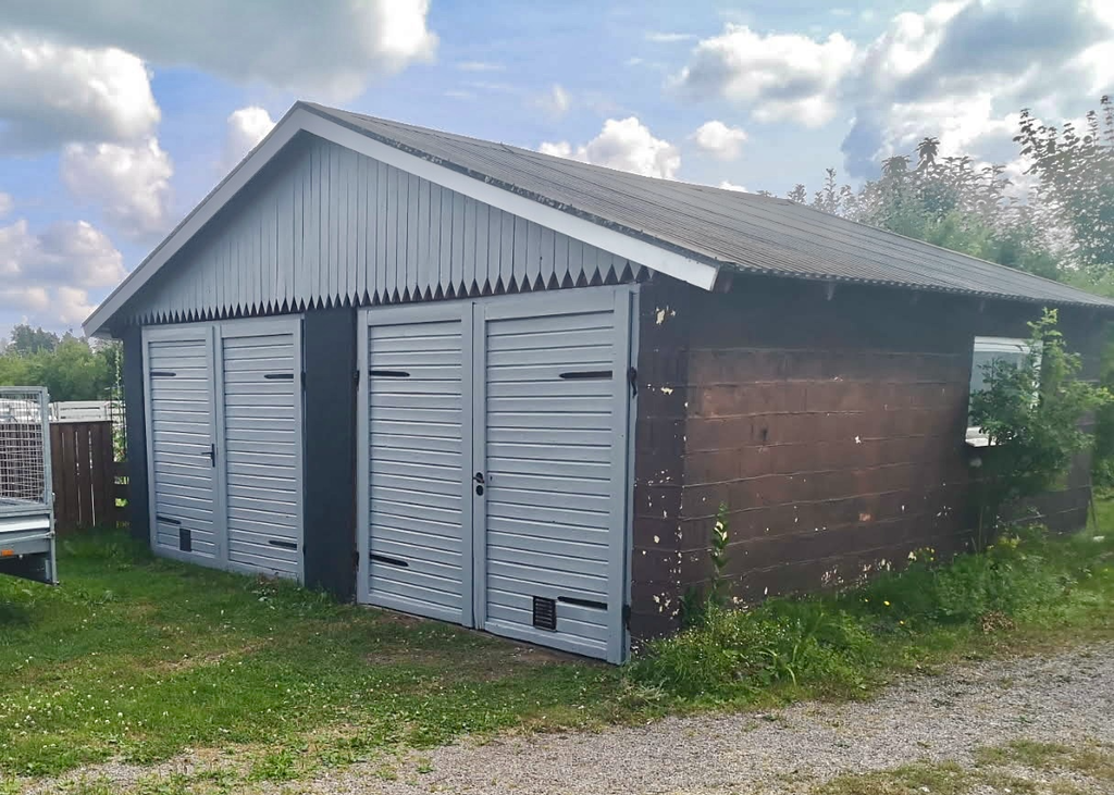 Enklare garage med grusat golv, fasad av panel (målad 2025) och plåttak från ca 2015. Garaget delas med grannen.