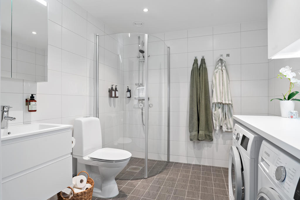 Badrummet går i vitt, med grått kakel på golv. Tvättavdelning med tvättmaskin och torktumlare från Siemens. Bänkyta ovanpå för tvätthantering samt förvaring