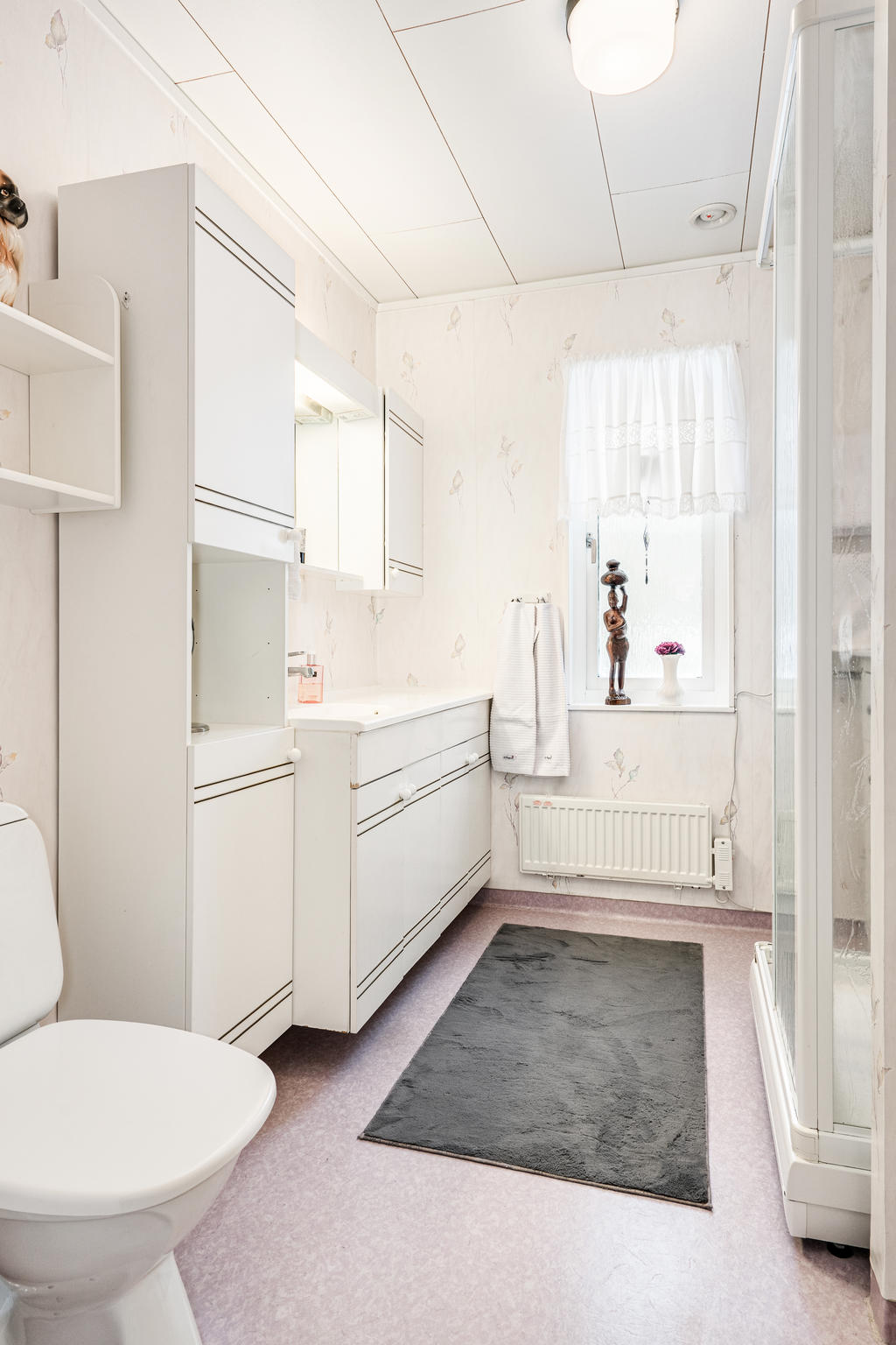 Ljust badrum med duschkabin, toalett och handfat med kommod. 