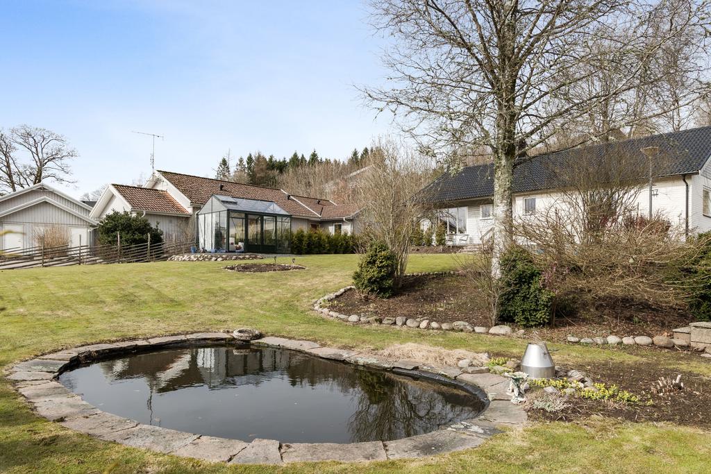 En lugn och välskött trädgård med damm i förgrunden, omgiven av grönska och blomrabatter.