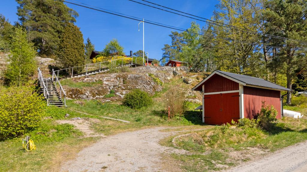 Parkering nere vid grusvägen med carport (med enkla dörrar) och trappor upp till huset