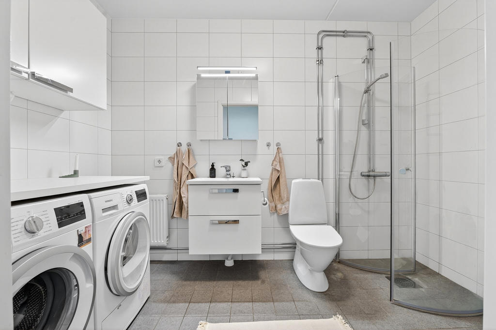 Stort badrum utrustat med wc, handfat, badrumsskåp, dusch samt tvättmaskin och torktumlare med arbetsbänk och förvaring.