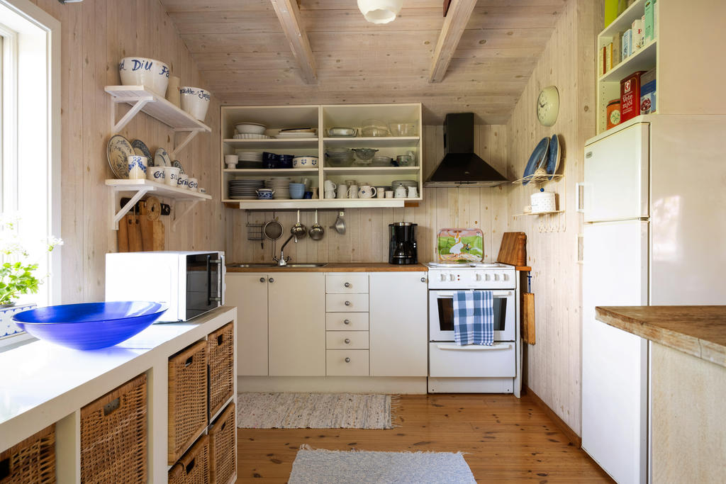 Det charmiga köket präglas av en varm och inbjudande atmosfär med ljusa träpaneler på väggar och i tak som skapar en genuin stugkänsla. Här finns plats för förvaring i både skåp och öppna hyllor som ger köket karaktär och plats för vackert porslin och köksdetaljer. Arbetsytorna i trä bidrar till den hemtrevliga känslan och tillsammans med fönstret som släpper in rikligt med dagsljus skapas en trivsam miljö för matlagning och umgänge.