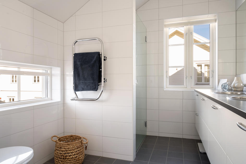 Stilrent och helkaklat badrum med vita kaklade väggar och mörkgrått klinkergolv. Badrummet är utrustat med duschhörna med glasvägg, wc, handfat med kommod och goda förvaringsmöjligheter. Fönster ger ett behagligt naturligt ljusinsläpp och den vägghängda handdukstorken bidrar till extra komfort.